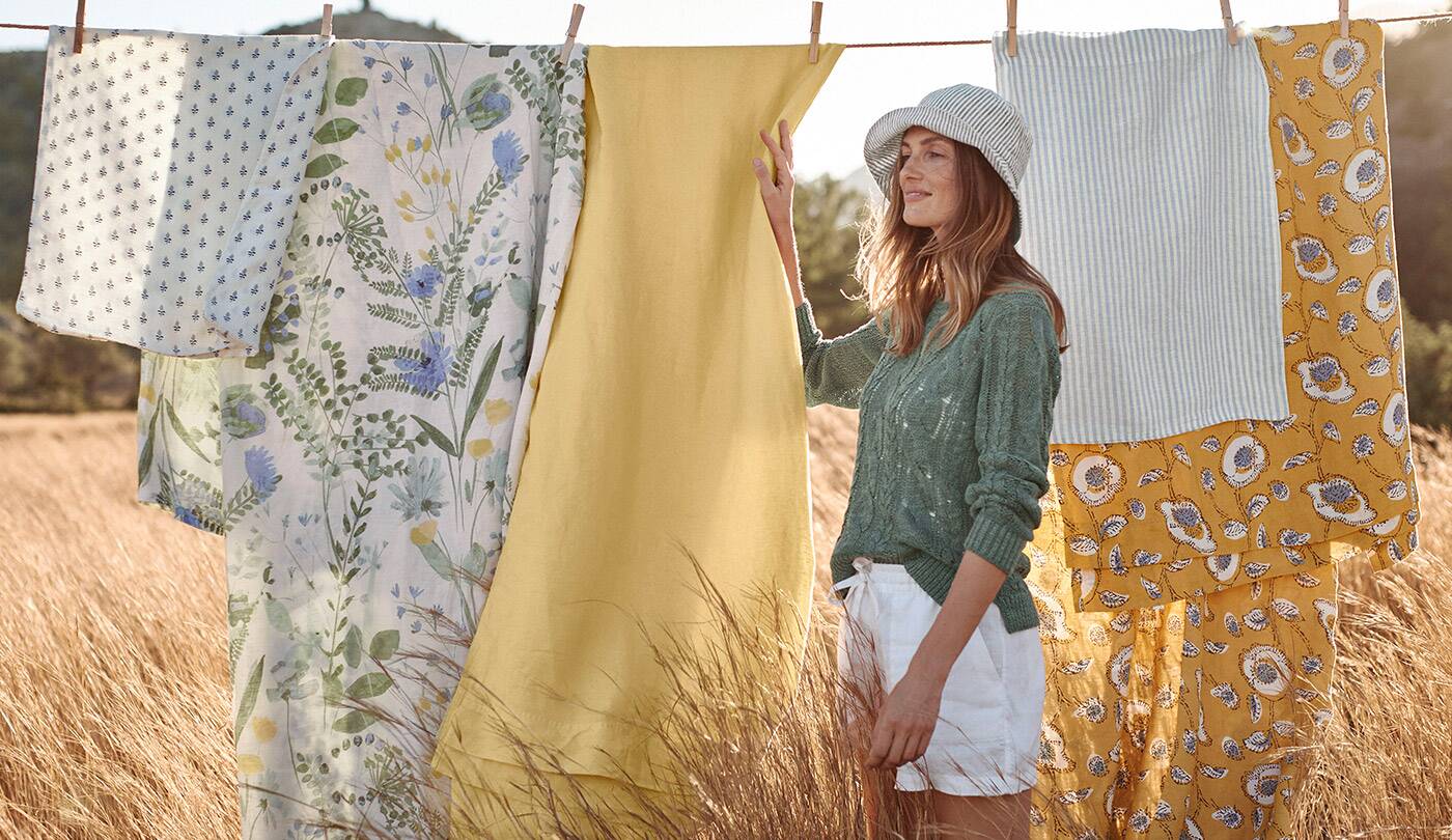 A woman in a green sweater stands at a clothesline with linen bedding. Browse women's clothing.